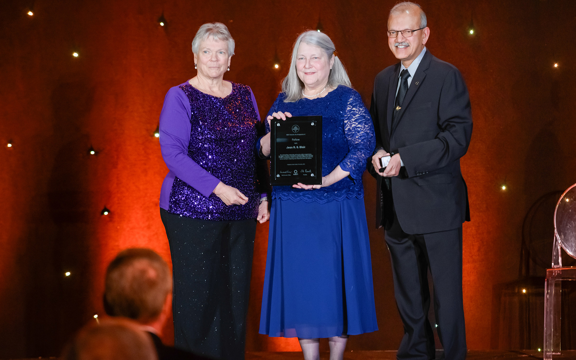 Two women and an older man standing on stage while the woman in the middle holds an award